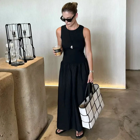 Woman in a black dress holding a white tote bag and a drink, standing in a modern indoor setting.